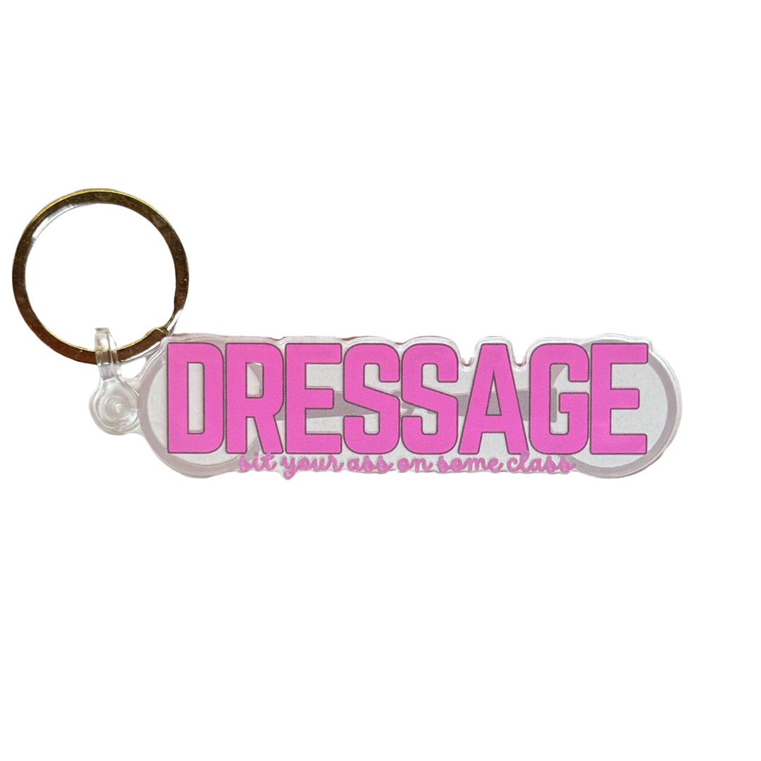 Dressage keychain. Large pink text reads “dressage” with smaller text “sit your ass on some class” over the background image of a loose ring snaffle bit.