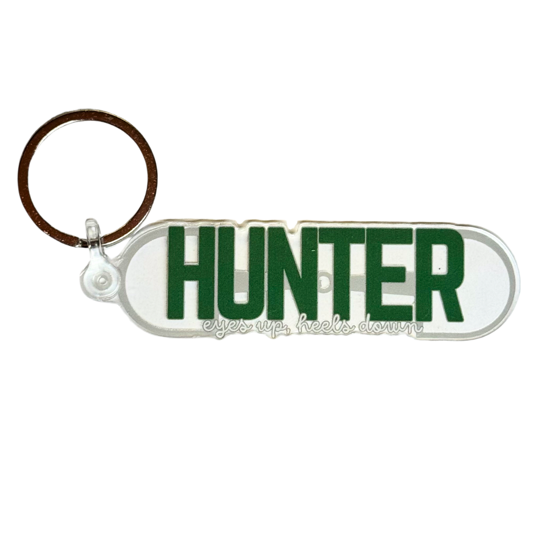 Hunter horse keychain. Large green text reads “hunter” with smaller text “eyes up, heels down” over background image of a dee ring snaffle bit.